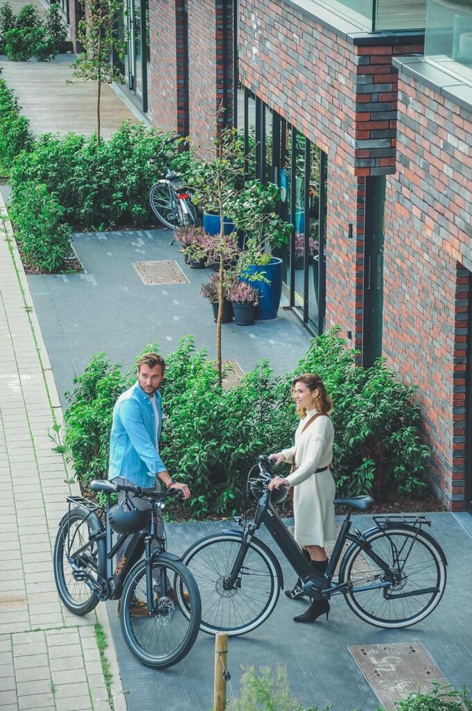 A man and a woman standing by their electric bikes in front of a brick building